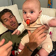 Sienna is registered to the contest to win money with this photo: baby, man, carrot, couch, tattoo, socks, hand, indoor, person, food, fruit, clothing, facial_expression, home, candid, holding, cute, infant, beige_outfit, relaxed