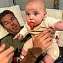 baby, man, carrot, couch, tattoo, socks, hand, indoor, person, food, fruit, clothing, facial_expression, home, candid, holding, cute, infant, beige_outfit, relaxed