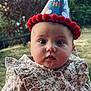 baby, child, birthday_hat, pom_poms, floral_dress, ruffles, portrait, face, wide_eyes, outdoor, grass, nature, celebration, cute, infant, young_child, close_up, person, headwear, festive