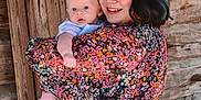 Jimmy is registered to the contest to win money with this photo: woman, baby, child, floral_dress, smile, portrait, outdoor, wooden_wall, holding, person, cute, infant, happy, family, love, casual, natural_light, skin, head, face