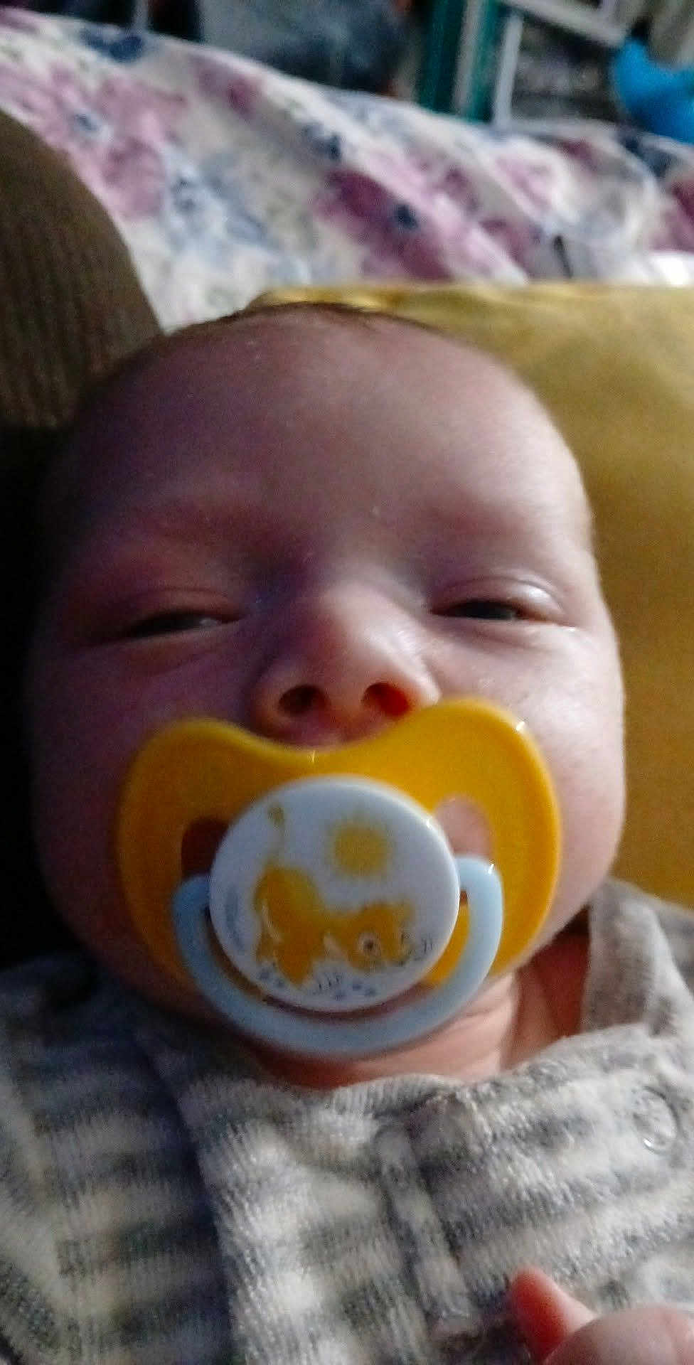 Phoenix is registered to the contest to win money with this photo: baby, pacifier, close_up, face, infant, sleepy, yellow, lion, clothing, striped, hand, indoors, portrait, child, young, cute, cozy, comfort, soft_focus, home