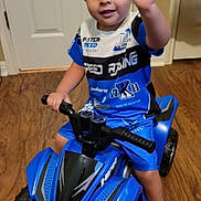 Jasaya is registered to the contest to win money with this photo: child, toddler, boy, smiling, waving, portrait, indoor, toy_vehicle, quad_bike, ride_on_toy, blue, shirt, shorts, boots, helmetless, wooden_floor, door, living_room, vehicle, happy