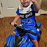 child, toddler, boy, smiling, waving, portrait, indoor, toy_vehicle, quad_bike, ride_on_toy, blue, shirt, shorts, boots, helmetless, wooden_floor, door, living_room, vehicle, happy