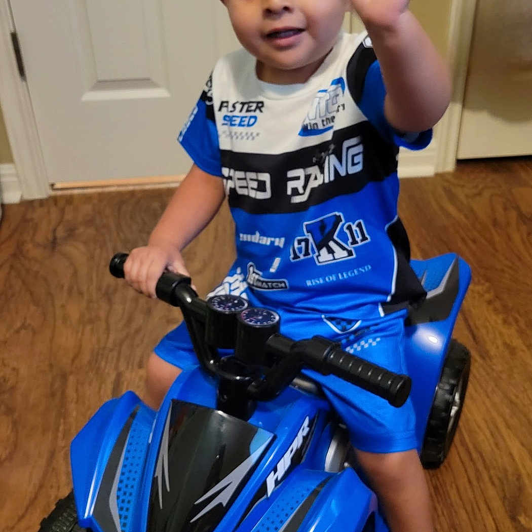 Jasaya is registered to the contest to win money with this photo: child, toddler, boy, smiling, waving, portrait, indoor, toy_vehicle, quad_bike, ride_on_toy, blue, shirt, shorts, boots, helmetless, wooden_floor, door, living_room, vehicle, happy