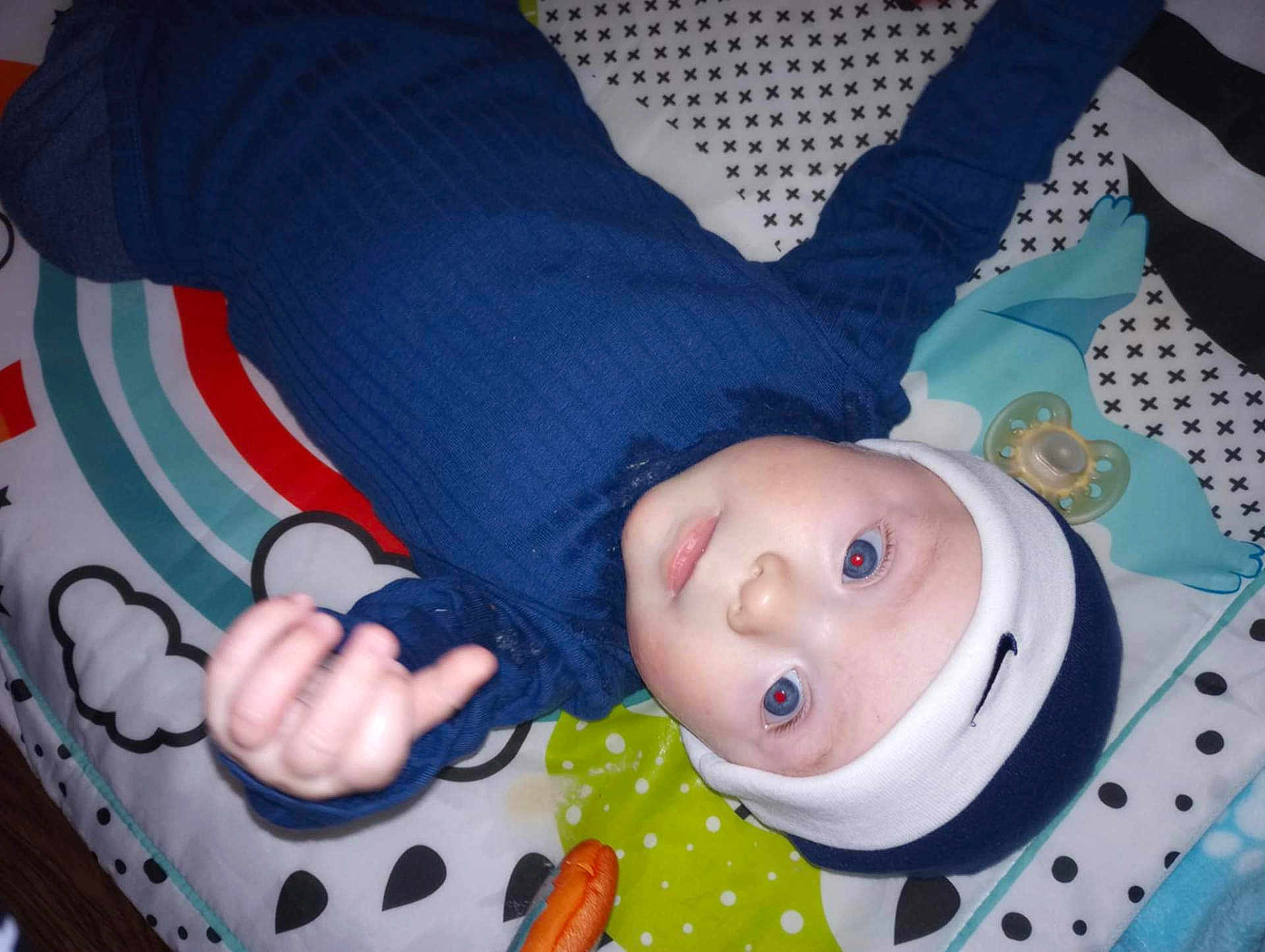 Theodore is registered to the contest to win money with this photo: baby, child, blue_clothing, beanie, blanket, colorful, pattern, indoor, infant, laying_down, pacifier, person, portrait, reaching_hand, red_eye, soft_toys, toy, white, young_child, cute
