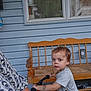 adult_partial, bench, bike, blue_siding, child, curious, daylight, handlebars, home, outdoor, patterned_shirt, person, porch, serious_expression, short_sleeve_shirt, toddler, toy, window, wooden_bench, yellow_shorts