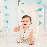 Bryan joined the competition — help win amazing prizes! baby, child, crawling, white_outfit, blue_eyes, balloons, neon_lights, star_shape, paper_airplanes, celebration, indoor, smiling, cute, decorations, floor_reflection, portrait, happy, light_background, infant, studio_photo