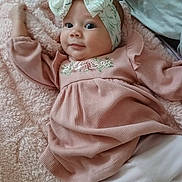 Alina is registered to the contest to win money with this photo: baby, infant, headband, pink_dress, blanket, smile, face, cute, child, lying_down, cozy, soft_texture, indoor, portrait, young_child, apparel, headwear, happy, closeup, person