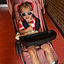 Harlo is registered to the contest to win money with this photo: toddler, child, stroller, sunglasses, toy, pink, red_bows, sitting, indoor, brick_wall, floor, black_shoes, white_socks, casual_clothing, smiling, person, young_child, playful, comfort, baby_stroller