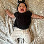 Alora-Mae is registered to the contest to win money with this photo: baby, smiling, black_bow, headband, black_top, white_pants, knit_pants, socks, bedspread, patterned_bedspread, lying_down, happy, cheerful, infant, child, cute, indoors, portrait, person, playful