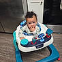child, baby, infant, baby_walker, toy, kitchen, hardwood_floor, refrigerator, cabinet, oven, curly_hair, hand_in_mouth, teething, sitting, looking_at_camera, curious, portrait, play_tray, colorful_toys, floor
