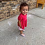 toddler, child, outdoor, sidewalk, chalk_drawings, brick_wall, red_top, patterned_shorts, shoes, curly_hair, smiling, standing, concrete, planter, window, casual_clothing, young_child, daylight, playtime, earrings