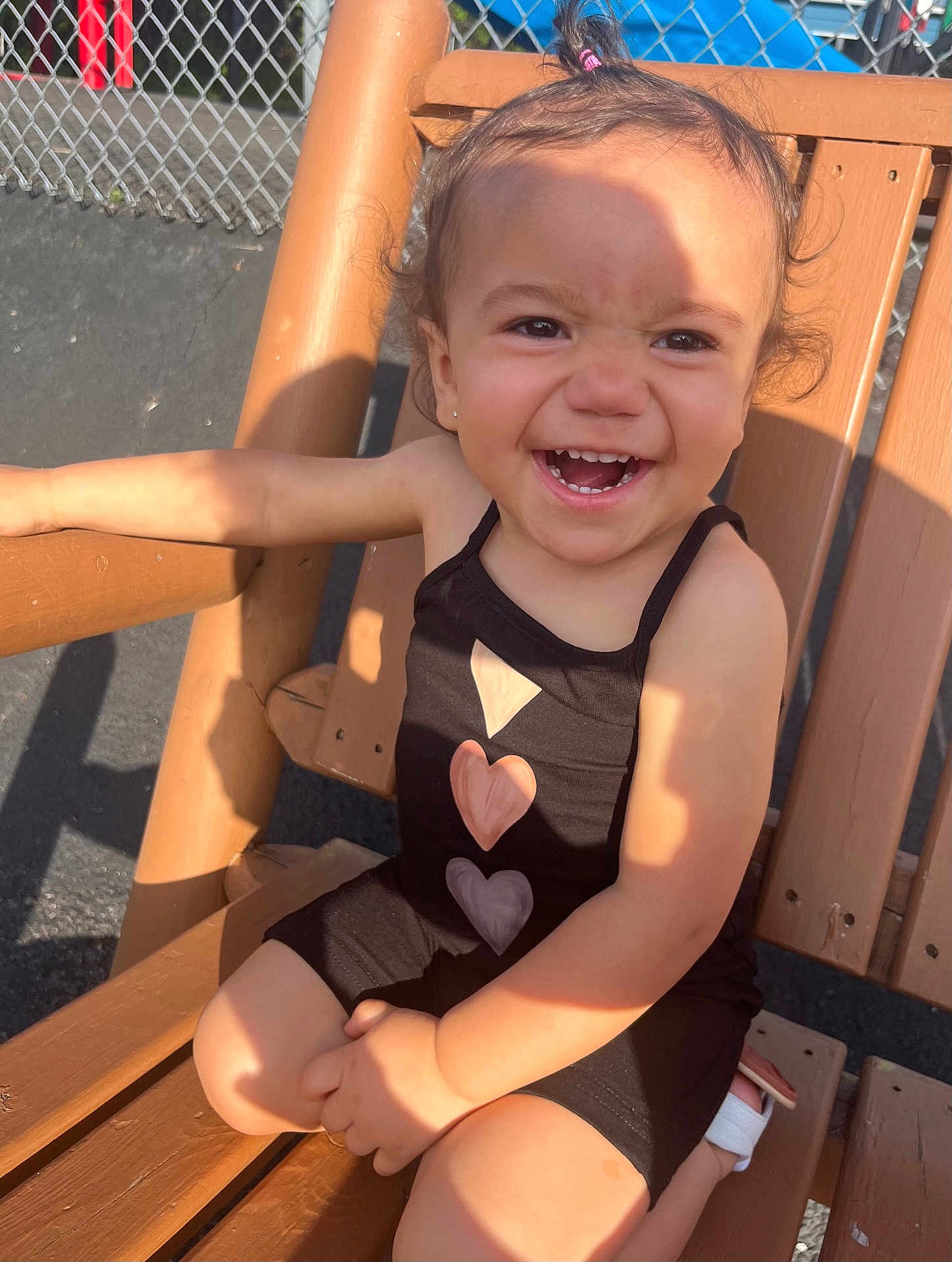 Ella is registered to the contest to win money with this photo: toddler, child, smiling, outdoor, bench, wooden_bench, black_outfit, heart_pattern, sunlight, shadow, happy, cute, person, summer, playful, hair_tie, casual, daytime, expression, skin