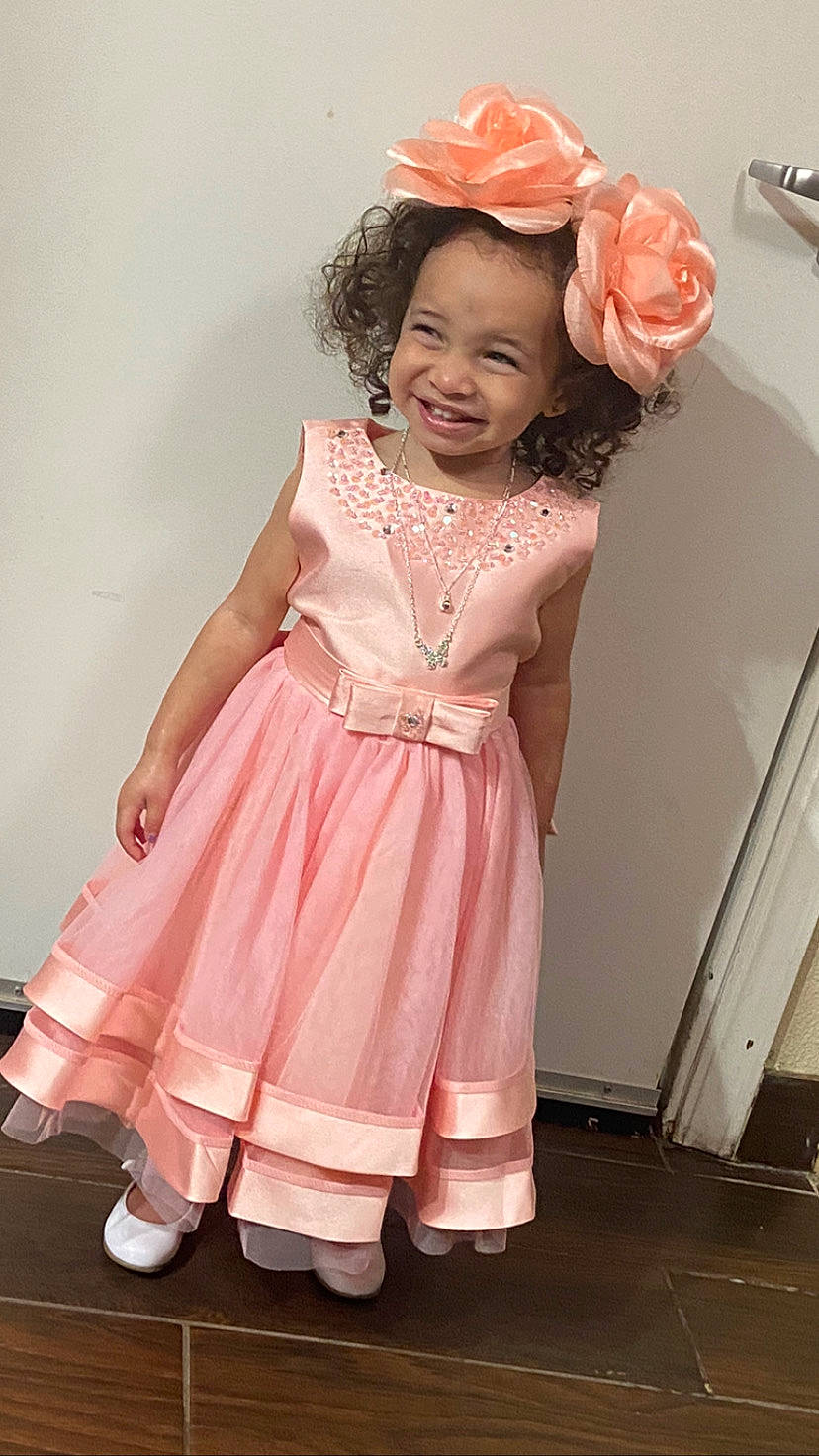 Danielle is registered to the contest to win money with this photo: baby_toddler_clothing, day_dress, dress, embellishment, fashion_design, flower, happy, hat, headpiece, joy, magenta, one_piece_garment, person, pink, rose, ruffle, skin, sleeve, smile, standing
