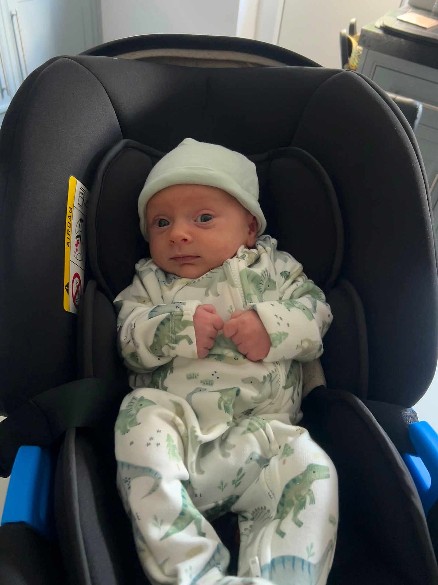 Noah joined the competition — help win amazing prizes! baby, infant, car_seat, onesie, hat, dinosaur_print, clothing, child, young, indoor, seat, fist, cute, newborn, person, portrait, sitting, black, soft_lighting, expression