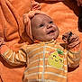 baby, child, headband, mittens, blanket, orange, smile, face, person, infant, cute, striped_clothing, zipped_onesie, soft_texture, happy, lying_down, closeup, cozy, interaction, sibling