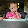 toddler, child, high_chair, pink_shirt, food, eating, indoor, face, hand, table, ceiling_fan, plant, furniture, window, curtains, person, portrait, baby, room, home