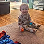 toddler, child, pajamas, football_pattern, rug, floor, balls, smiling, indoors, blanket, storage_drawers, wooden_floor, toy, happy, person, home, living_room, casual, clothing, young_child