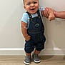 toddler, child, smiling, denim_overalls, gray_sneakers, standing, hand_support, indoor, wooden_floor, white_wall, cute, happy, baby, person, footwear, casual_clothing, young_child, portrait, human, one_person