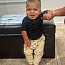 Mateo joined the competition — help win amazing prizes! baby, child, indoor, standing, smiling, black_shirt, cream_pants, barefoot, hand_support, carpet, ottoman, window, person, cute, happy, home, human, infant, young_child, light