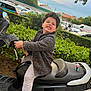 black_scooter, cars, casual_clothing, child, cloudy, curly_hair, funny_face, greenery, grimace, grip, hedge, jacket, outdoor, pants, parking_lot, scooter, shoes, sky, toy_figure, urban