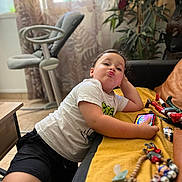 Mylann participe au concours pour gagner de l'argent avec cette photo : child, indoor, playing, smartphone, toys, pacifier, couch, blanket, yellow, white_shirt, black_shorts, floor, face, funny_expression, plant, high_chair, table, relaxed, home, person