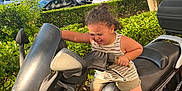 Mylann a rejoint le concours — aidez-le/la à gagner de superbes lots ! black_scooter, blue_crocs, car, child, curly_hair, daylight, determined, greenery, handlebars, hedge, outdoor, parking_lot, play, scooter, shorts, sunlight, tank_top, toy, tree, vehicle