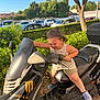 child, scooter, outdoor, vehicle, greenery, tree, sunlight, hedge, parking_lot, car, toy, blue_crocs, shorts, tank_top, curly_hair, determined, play, handlebars, black_scooter, daylight