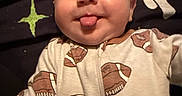 Zayne is registered to the contest to win money with this photo: baby, infant, face, tongue, onesie, clothing, football, pattern, blanket, lying_down, cheeky, cute, smile, child, person, head, skin, indoors, sleepwear, expression