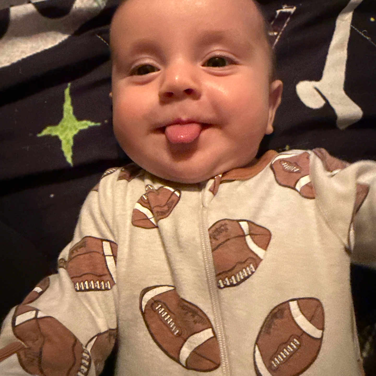 Zayne is registered to the contest to win money with this photo: baby, infant, face, tongue, onesie, clothing, football, pattern, blanket, lying_down, cheeky, cute, smile, child, person, head, skin, indoors, sleepwear, expression