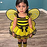 child, girl, bee_costume, yellow, black, antennae, wings, dress, tutu, striped_socks, striped_shoes, flowers, portrait, standing, indoors, wood_floor, blue_wall, costume, fashion, smiling