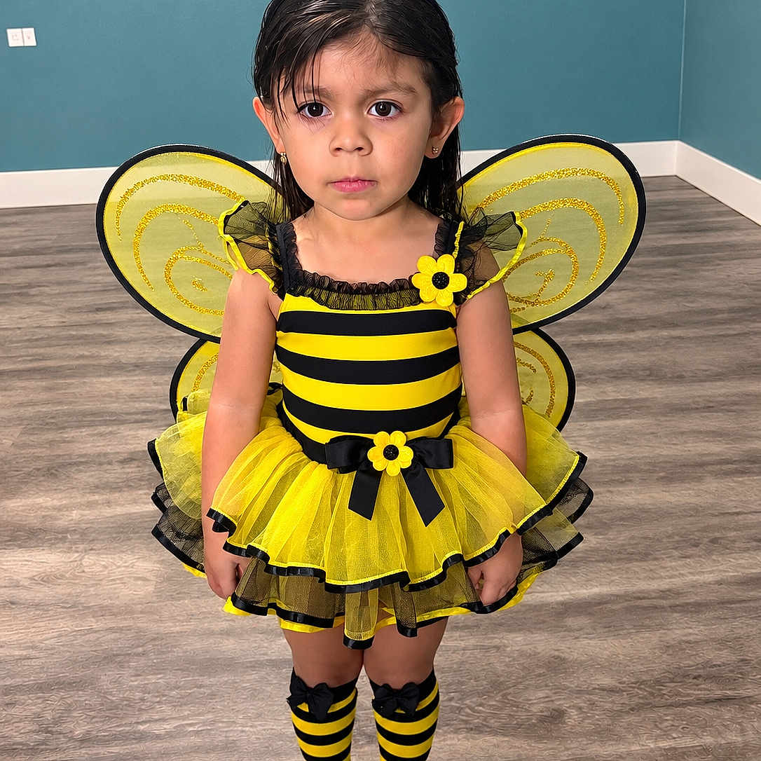 Carolina Vasquez joined the competition — help win amazing prizes! child, girl, bee_costume, yellow, black, antennae, wings, dress, tutu, striped_socks, striped_shoes, flowers, portrait, standing, indoors, wood_floor, blue_wall, costume, fashion, smiling