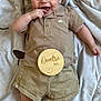 baby, infant, smiling, clothing, polo_shirt, shorts, blanket, wooden_sign, lying_down, hand, foot, face, cute, child, indoors, soft_texture, happy, skin, young, portrait