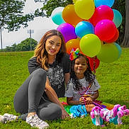 Mia is registered to the contest to win money with this photo: balloons, casual_clothing, child, colorful, daytime, fun, grass, greenery, happy, leisure, nature, outdoor, park, people, playful, roller_skates, sitting, smiling, tree, woman