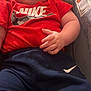 arm, baby, blue_pants, brand_logo, car_seat, child, chubby_cheeks, closeup, clothing, cushion, expressionless, hand, indoors, infant, nike_logo, nike_shirt, portrait, red_tshirt, sitting, soft_lighting