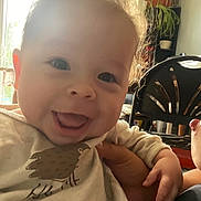 Milo is registered to the contest to win money with this photo: baby, smiling, curly_hair, indoor, child, person, happy, hand, clothing, window, plant, furniture, chair, natural_light, face, portrait, cute, expression, toddler, cozy