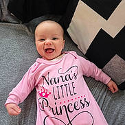 Kalilah is registered to the contest to win money with this photo: baby, infant, smiling, onesie, pink_clothing, couch, pillow, geometric_pattern, happy, portrait, indoor, child, cute, face, person, lying_down, cozy, soft_fabric, home, newborn