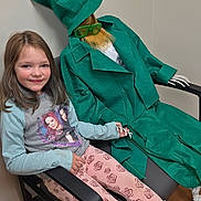 Harley is registered to the contest to win money with this photo: child, girl, smile, chair, costume, mannequin, skeleton_hand, green_hat, green_coat, sunglasses, beard, indoor, portrait, leggings, shoes, armrest, hair, playful, waiting_room, fabric