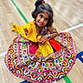 child, girl, smiling, traditional_clothing, colorful_dress, patterned_fabric, sitting, wooden_floor, sports_court_lines, hair_accessories, happy, portrait, indoor, cultural, festive, young, cute, posing, bright_colors, accessories