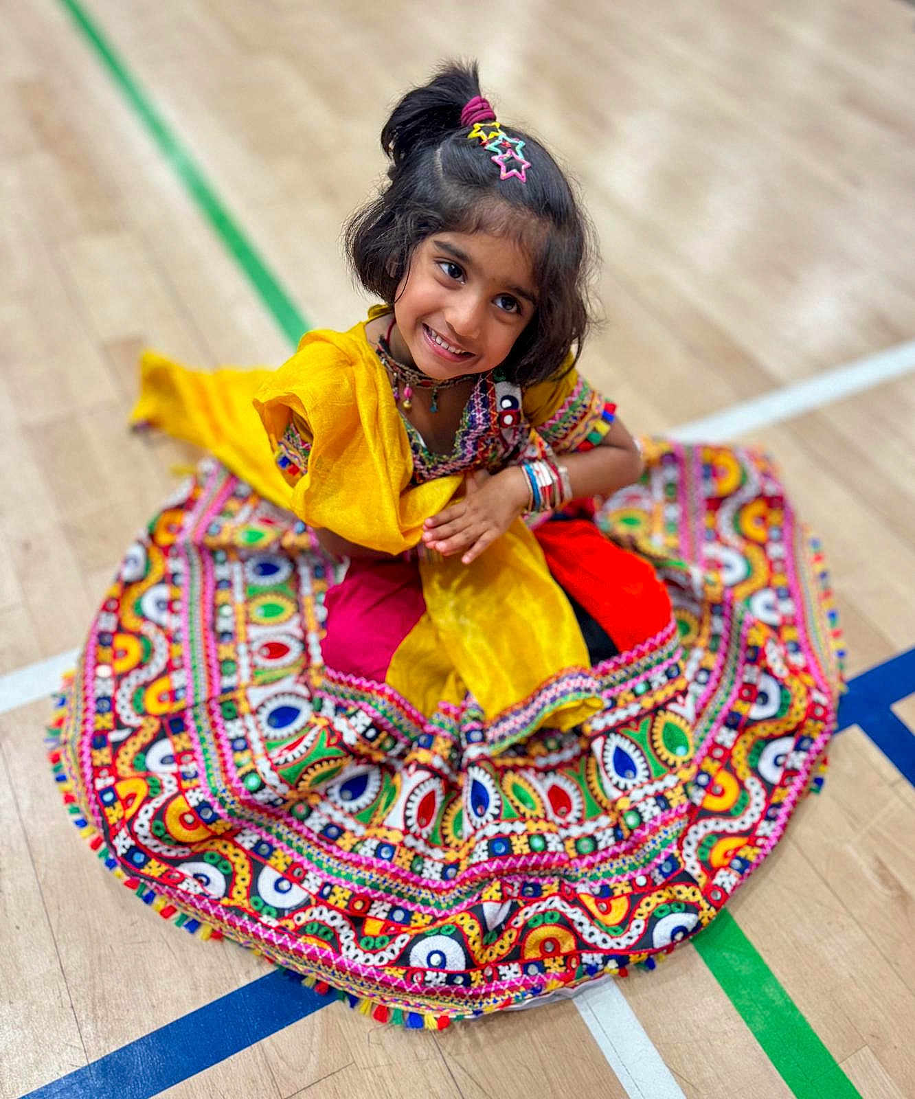 Qiyara joined the competition — help win amazing prizes! child, girl, smiling, traditional_clothing, colorful_dress, patterned_fabric, sitting, wooden_floor, sports_court_lines, hair_accessories, happy, portrait, indoor, cultural, festive, young, cute, posing, bright_colors, accessories
