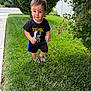 toddler, child, grass, lawn, tshirt, black_clothing, shorts, shoes, walking, outdoor, greenery, bush, leafy_plants, sidewalk, fence, suburban, portrait, young_child, nature, casual_clothing