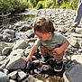 casual_clothing, child, creek, curiosity, daylight, exploration, footwear, greenery, hand, nature, outdoor, person, riverbed, rocks, shoes, squatting, sunlight, toddler, tool, water