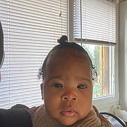 Jahëlya a rejoint le concours — aidez-le/la à gagner de superbes lots ! baby, child, face, portrait, indoors, window, blinds, natural_light, curly_hair, top_knot, bib, clothing, person, young_child, close_up, expression, skin, head, eye, cute