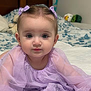 Ariana joined the competition — help win amazing prizes! baby, child, infant, purple_dress, bows, bed, blanket, wooden_headboard, plush_toys, indoor, portrait, face, eyes, cute, soft_lighting, curious, skin, hands, toddler, young_child