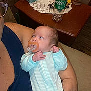 Trinity joined the competition — help win amazing prizes! baby, pacifier, adult, glasses, blue_clothing, arm, hand, table, bottle, sprite, doily, furniture, indoors, person, face, looking, holding, skin, resting, curious