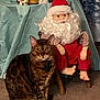 cat, indoor, decor, santa_doll, figurines, table, carpet, curtain, beard, red_clothing, striped_sleeves, brown, spotted, seated, furniture, decorative, holiday, animal, pet, cozy