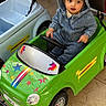 toddler, child, denim_hoodie, toy_car, green_car, unicorn_stickers, stars, indoor, floor_tiles, small_wheels, steering_wheel, cute_expression, casual_clothing, playtime, vehicle_toy, kid_playing, seat, hood, bright_colors, imaginative_play