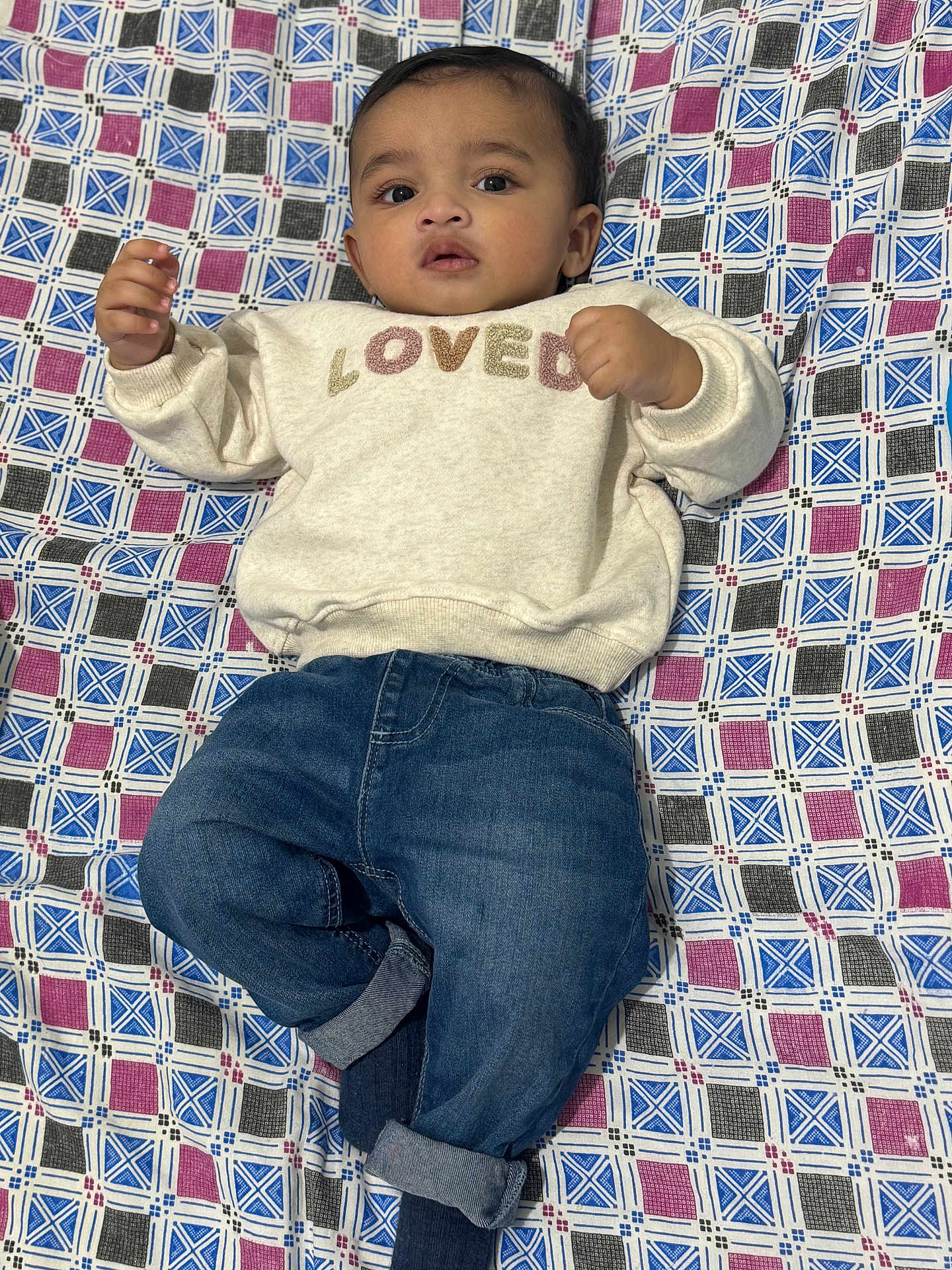 Saad is registered to the contest to win money with this photo: baby, child, infant, sweater, denim_jeans, blanket, patterned, lying_down, curious, indoors, portrait, casual_clothing, cute, young_child, face, hands, feet, relaxed, comfortable, closeup