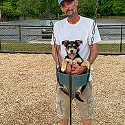 Toto is registered to the contest to win money with this photo: man, dog, swing, park, trees, wood_chips, cap, white_tshirt, shorts, outdoor, pet, happy, playground, fence, leash, smile, summer, casual_clothing, daytime, nature