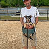 man, dog, swing, park, trees, wood_chips, cap, white_tshirt, shorts, outdoor, pet, happy, playground, fence, leash, smile, summer, casual_clothing, daytime, nature